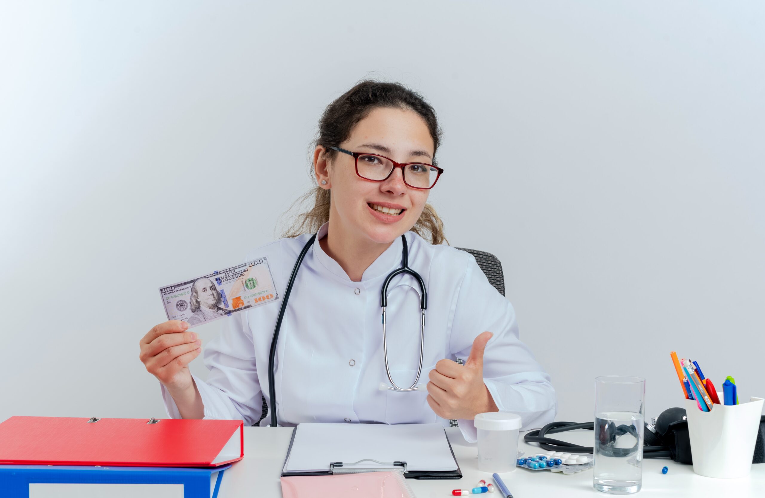 happy-young-female-doctor-wearing-medical-robe-stethoscope-glasses-sitting-desk-with-medical-tools-holding-money-looking-showing-thumb-up-isolated (1)